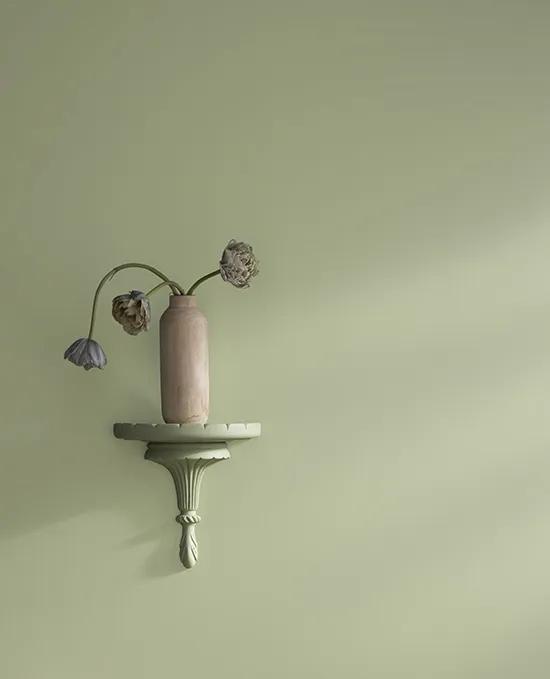 Sunlight reflects on a light green-painted wall and light-green carved wood wall sconce holding a vase of flowers.