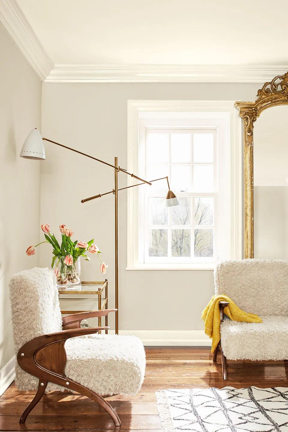 A warm gray living room features plush white Sherpa chairs, a modern floor lamp, and a large gilded mirror.