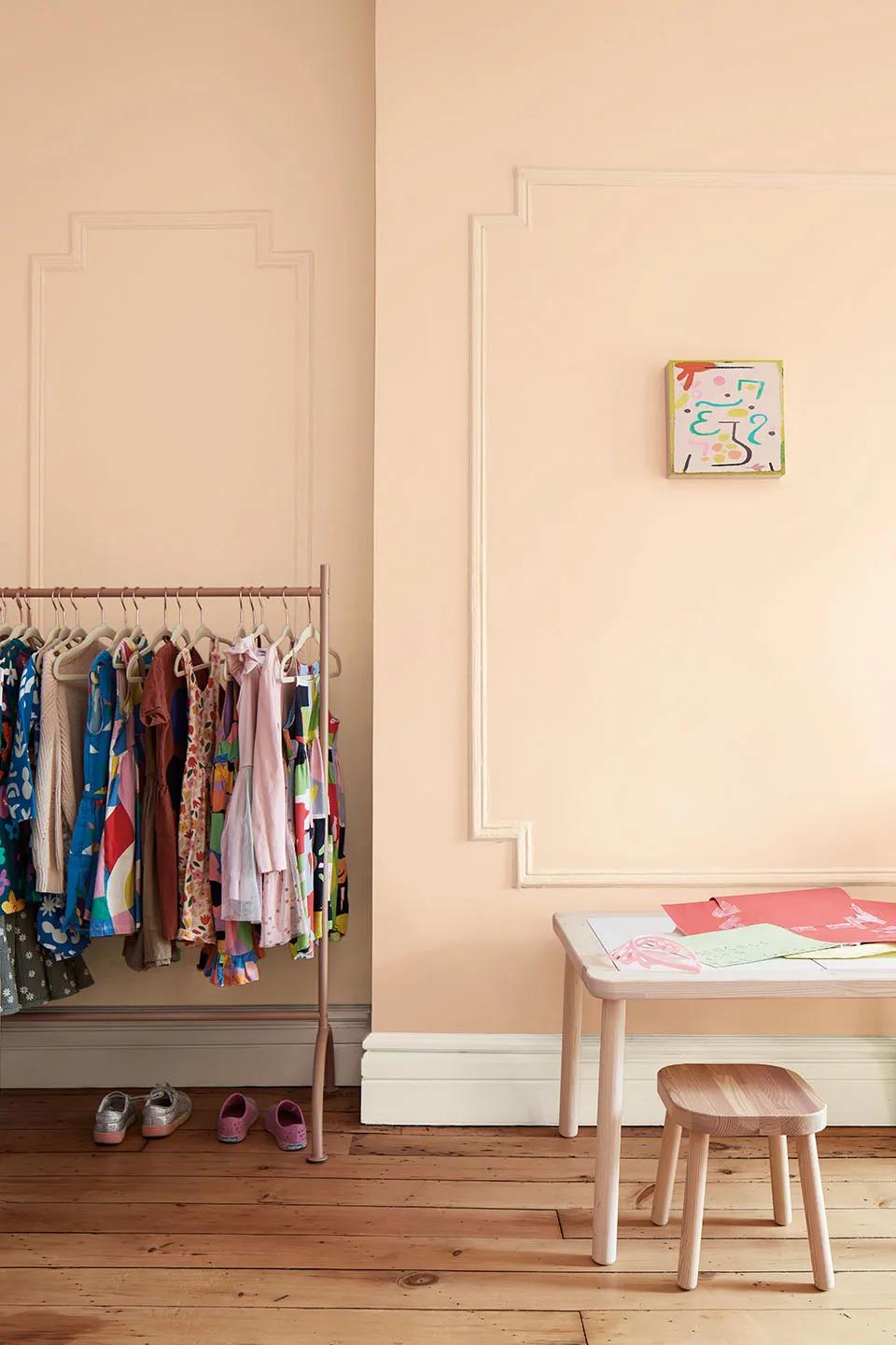 Una habitación de niño con paredes pintadas en rosa claro, una mesa de madera baja y un taburete, y un estante con vestimenta estampada.