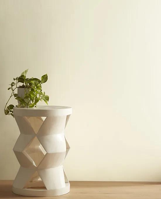 A white geometric shaped stool with a potted plant on top sits in front of a creamy, off-white painted wall.