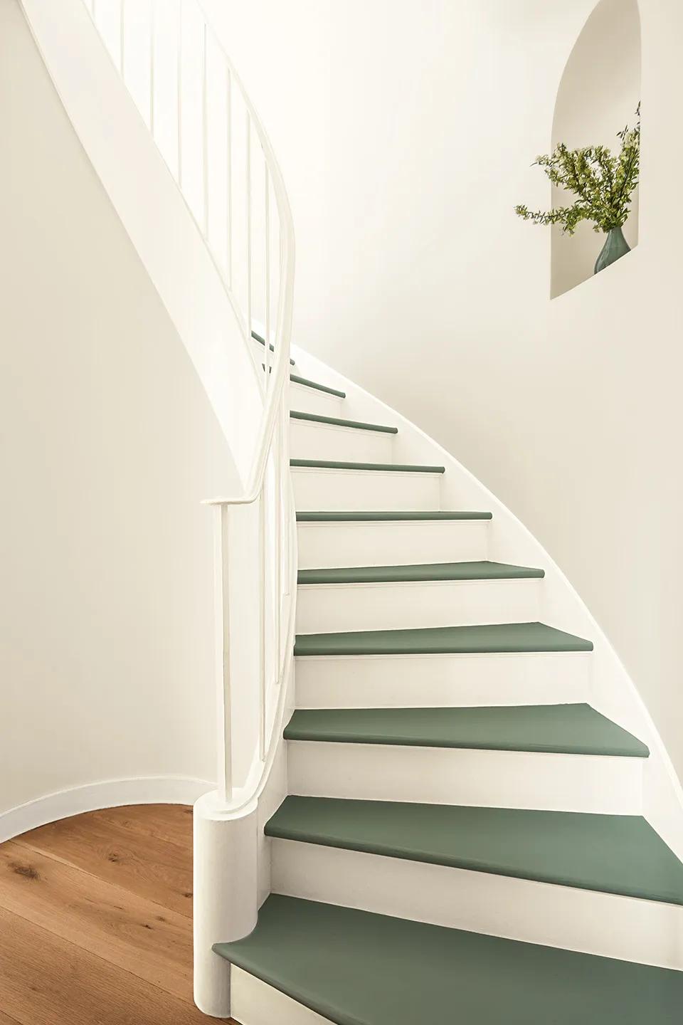 Una escalera de caracol en un pasillo blanco con contrahuellas pintadas de blanco y escalones pintados de verde.