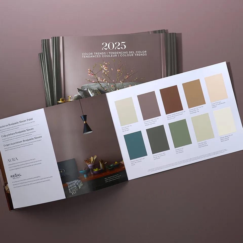 An open Benjamin Moore Colour of the Year 2025 brochure sits atop a stack of more brochures on a purple background.