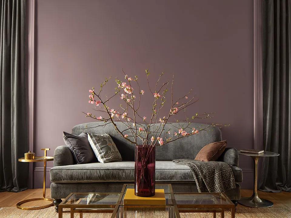 Una sala de estar pintada en púrpura con un sofá gris, mesas laterales doradas y una mesa de centro dorada con tapa de vidrio, con un jarrón rojo con flores rosas apoyado en ella.