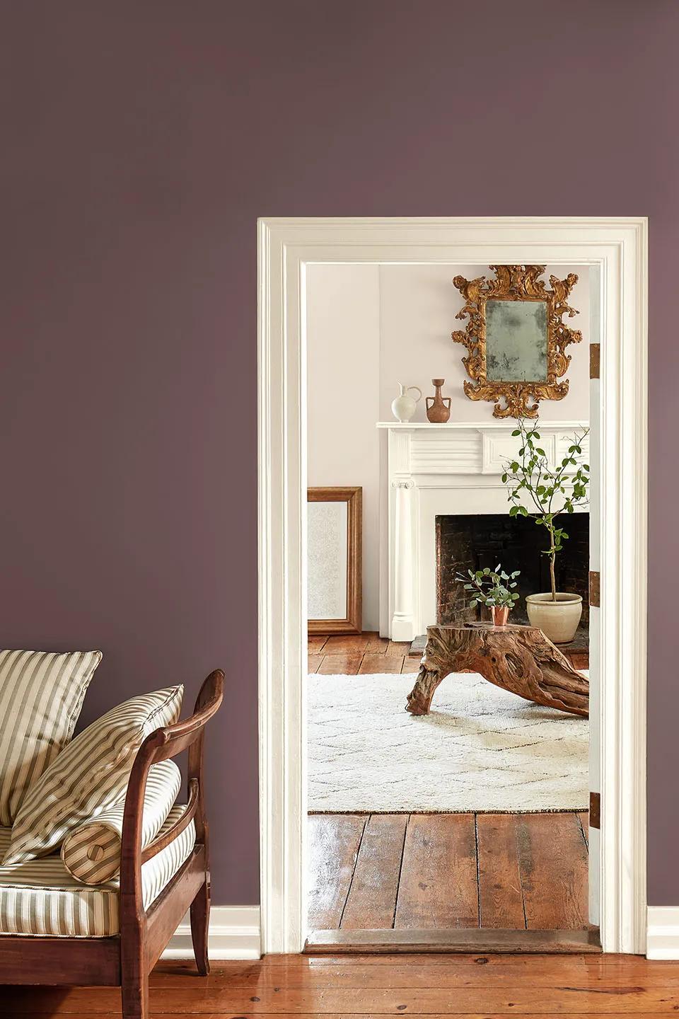 Una habitación pintada de púrpura con marco blanco y una silla de acento a rayas que conduce hacia una sala de estar pintada de blanco con una repisa, un espejo y detalles de madera.